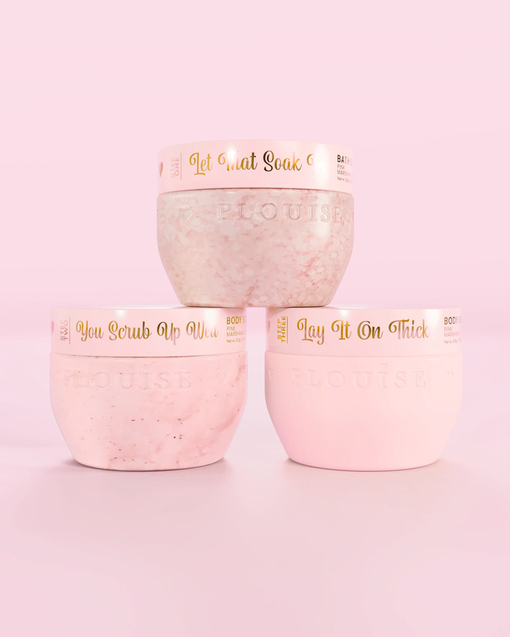 Tres recipientes rosas P.Louise Hop Into Self-Care Pink Marshmallow con sales de baño, exfoliantes corporales y manteca corporal con texto dorado sobre un fondo rosa