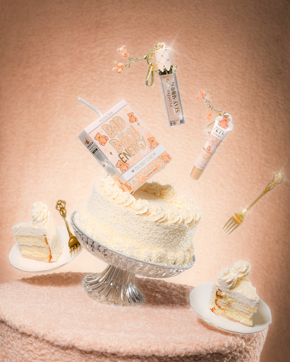 Un pastel sobre una mesa de tela de peluche con un dúo de labios P.Louise Teddy Glitter Bad Bitch Energy en pastel de coco que contiene un acondicionador de labios y aceite para labios flotando alrededor del pastel.