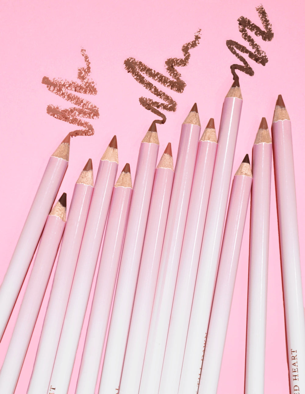 Ein Bild mehrerer gruppierter P.Louise Ultimate Definition Sharpenable Lip Liner, auf dem ein darüber gezeichnetes Produkt auf einem rosa Hintergrund zu sehen ist
