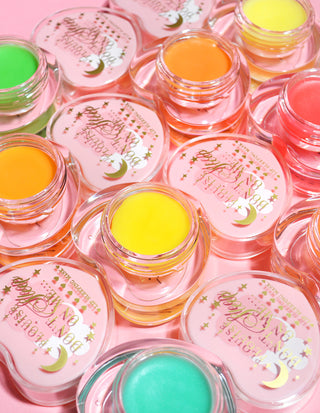 Multiple pots of lip sleeping mask with various colours including green, yellow, pink, and red, displayed on a pink background.