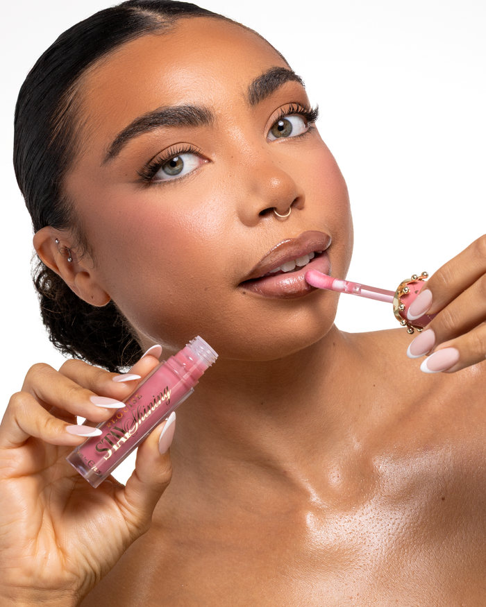 Eine Nahaufnahme einer Frau, die ein rosa P.Louise Stay Shining Lip Oil flüssiges Lippenprodukt mit einem Doe-Foot-Applikator aufträgt, während sie die Tube in der anderen Hand hält, Set vor einem sauberen weißen Hintergrund.