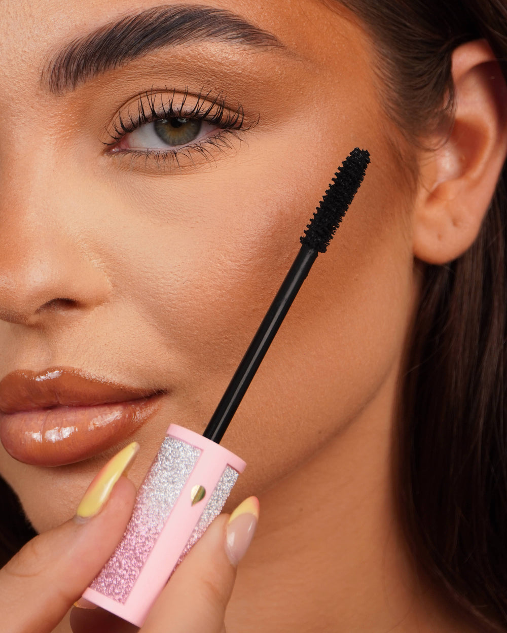Nahaufnahme eines Wimperntuschestabs von P.Louise Cry Baby, der mit einer rosa glitzernden Wimperntuschetube in die Nähe des Auges einer Person gehalten wird.