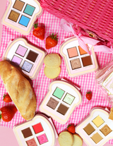 Un conjunto de mini paletas de sombras de ojos estilo sándwich con varios tonos de colores de maquillaje mostrados sobre una superficie de cuadros rosa, acompañados de fresas y un trozo de pan.