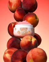 Glas mit P. Louise Slick It To Me Peach Melting Body Balm mit rosa Creme, umgeben von Pfirsichen auf einem Hintergrund mit Farbverlauf