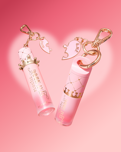 Zwei rosa Lipgloss-Röhren mit goldfarbenen Schlüsselanhänger-Clips und herzförmigen Anhängern, angezeigt auf einem zartrosa Hintergrund mit einer schwachen Herzkontur.