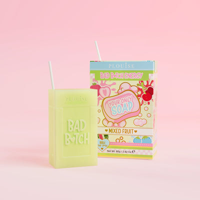 Una pastilla de jabón verde con la inscripción 'Bad Bitch' grabada, acompañada de una colorida caja de jugo con la etiqueta 'Mixed Fruit', presentada sobre un fondo rosa.