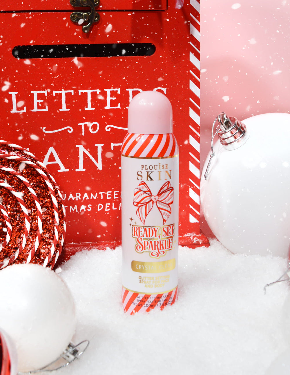 Eine Flasche P. Louise Ready Set Sparkle Christmas Glitter Setting Spray, umgeben von festlichen Dekorationen wie Schnee, Kugeln und einem Weihnachtsbriefkasten