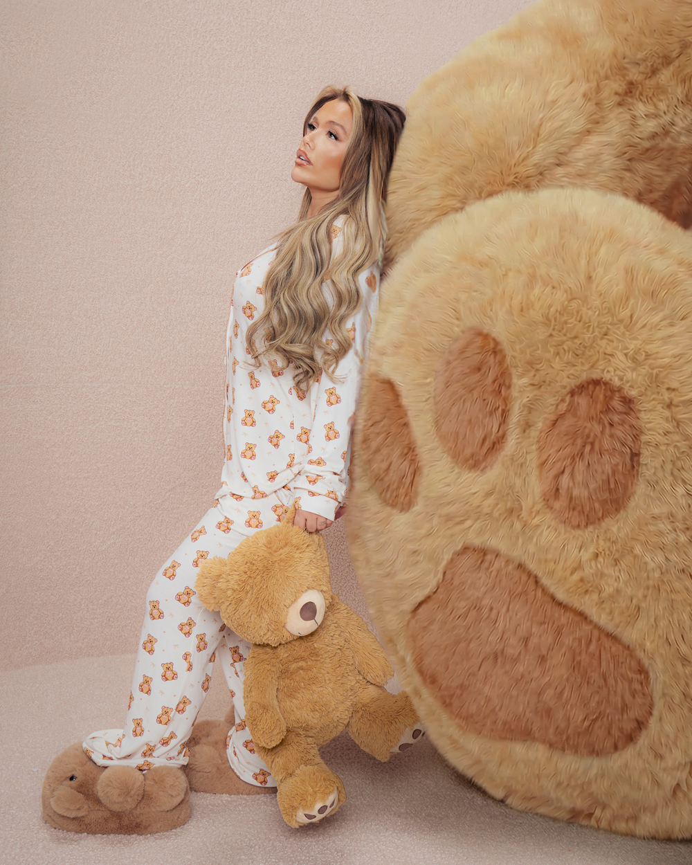 Mujer en pijama de peluche sosteniendo un osito de peluche junto a una gran pata de oso de peluche sobre un fondo beige