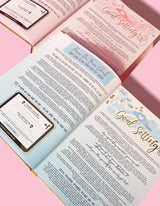 Un diario inspirador con varias páginas visibles, que muestra secciones sobre establecimiento de objetivos y otros temas de desarrollo, con un fondo rosa.